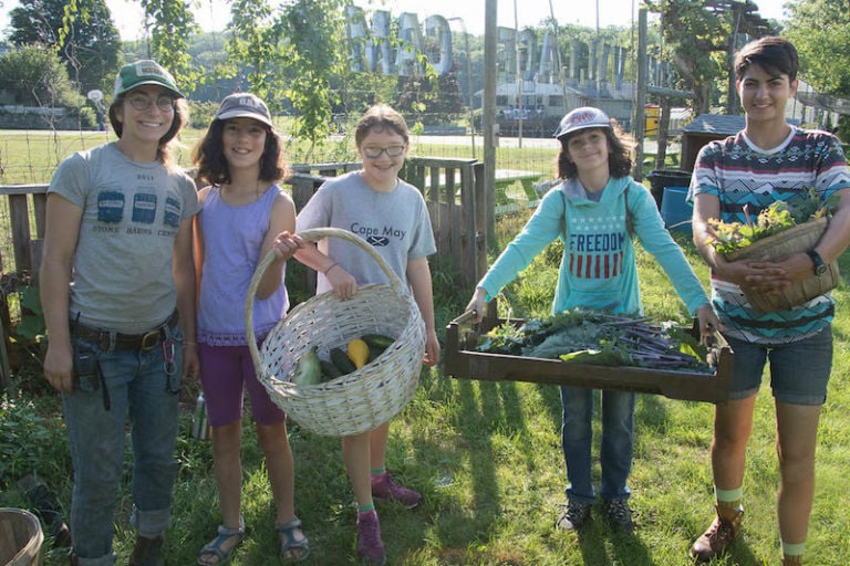 Five campers holding harvested vegetables in a sunny garden.