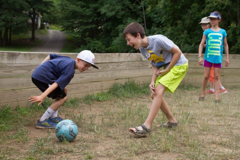 Children playing a ball game in a wooden enclosure outdoors.