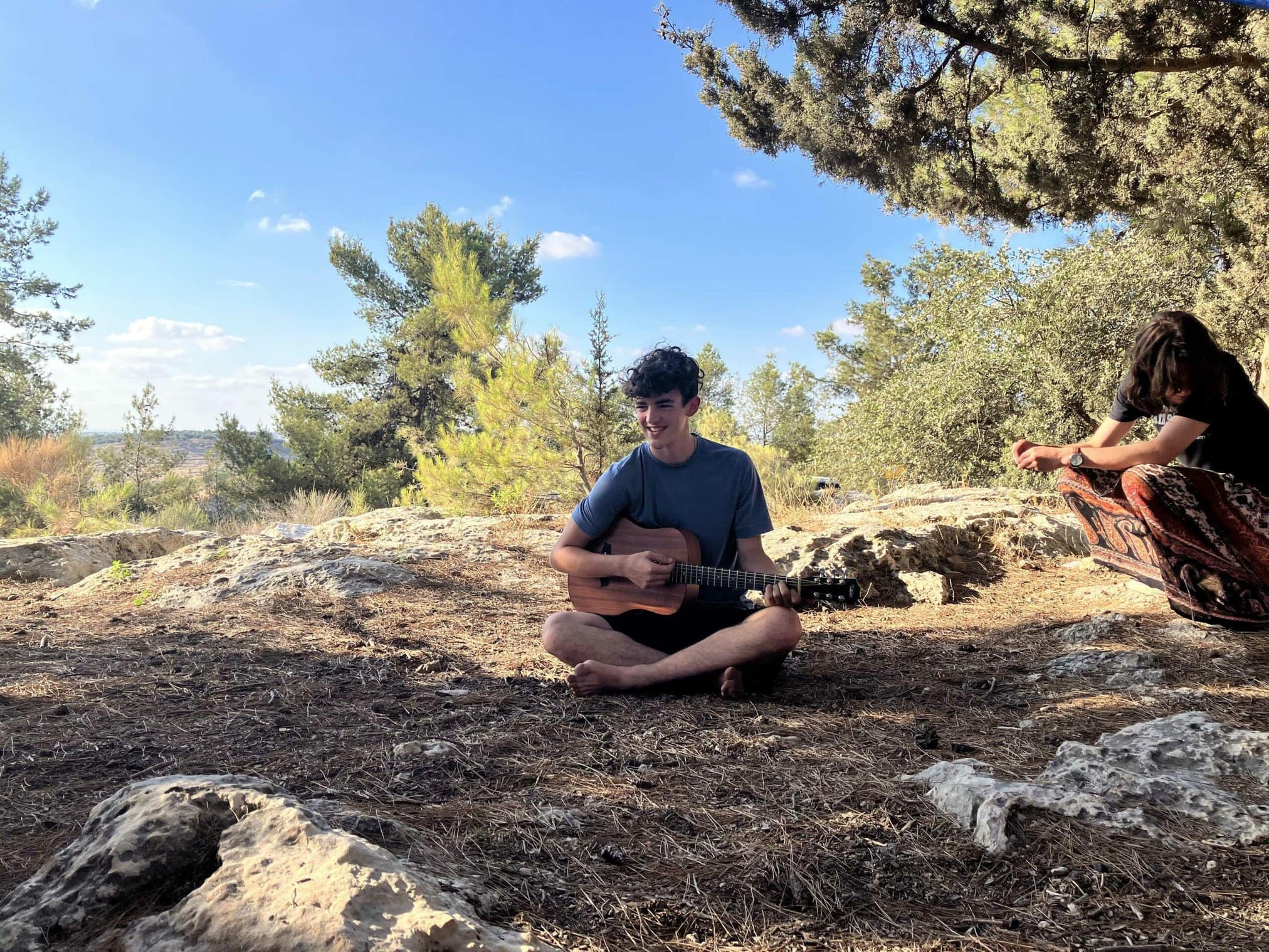 A person playing guitar outdoors with another individual seated nearby.