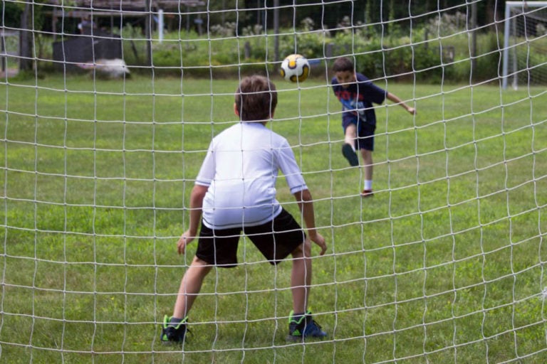 Two children playing soccer, one kicking the ball towards the goal.