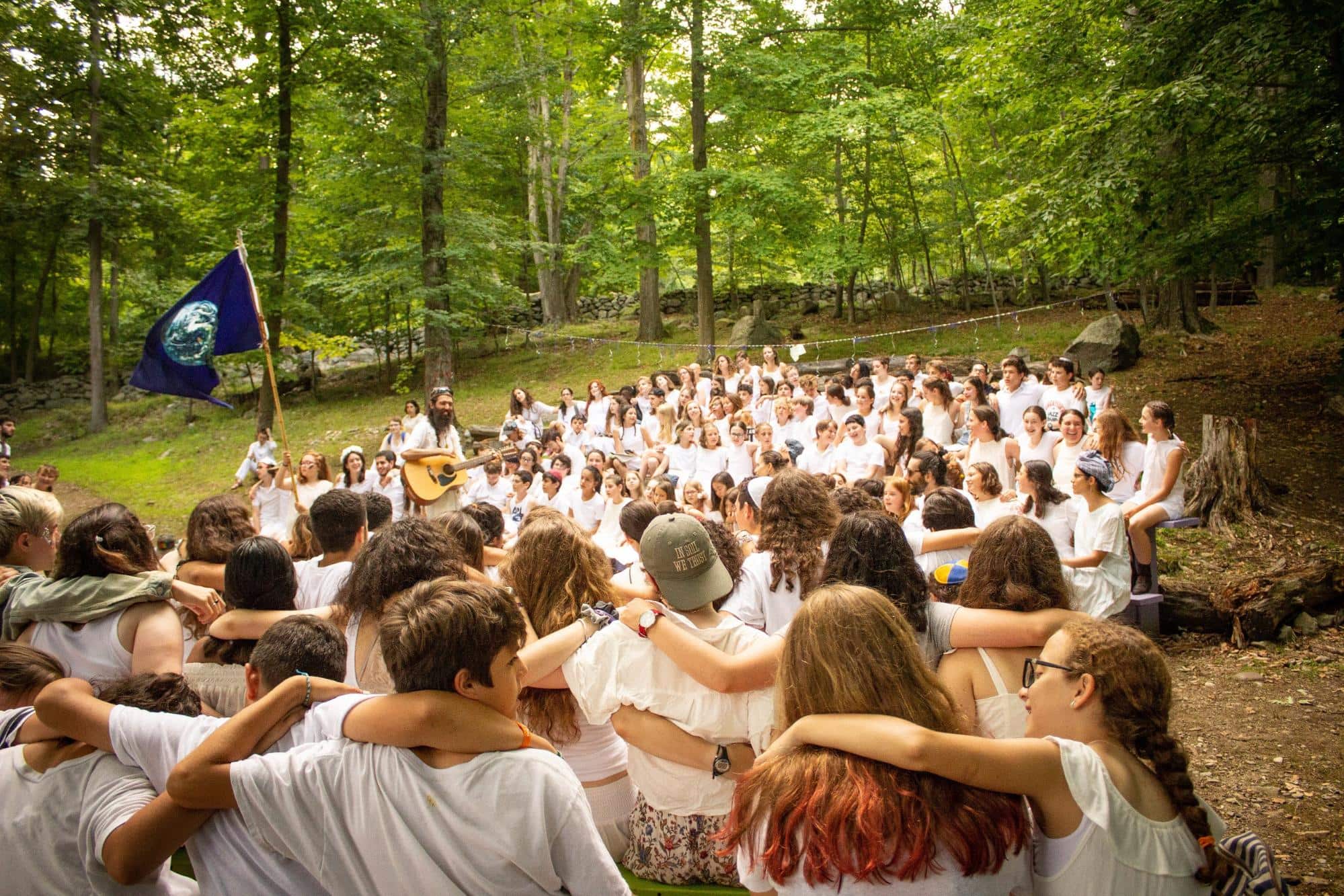 Large group gathered outdoors, embracing and singing in a forest setting.