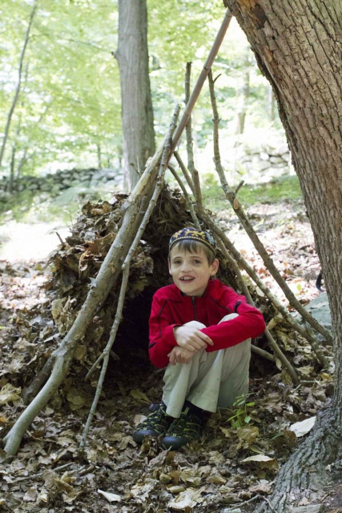 Child in red sweater sitting next to woodland stick shelter.