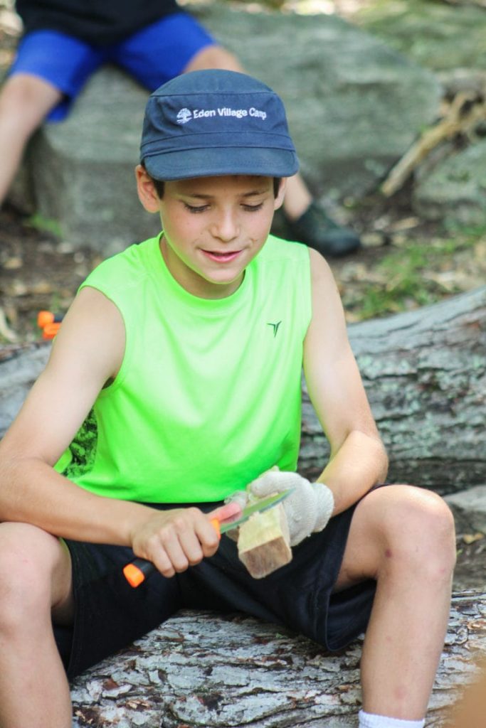 Boy in green shirt whittling wood outdoors on a sunny day.